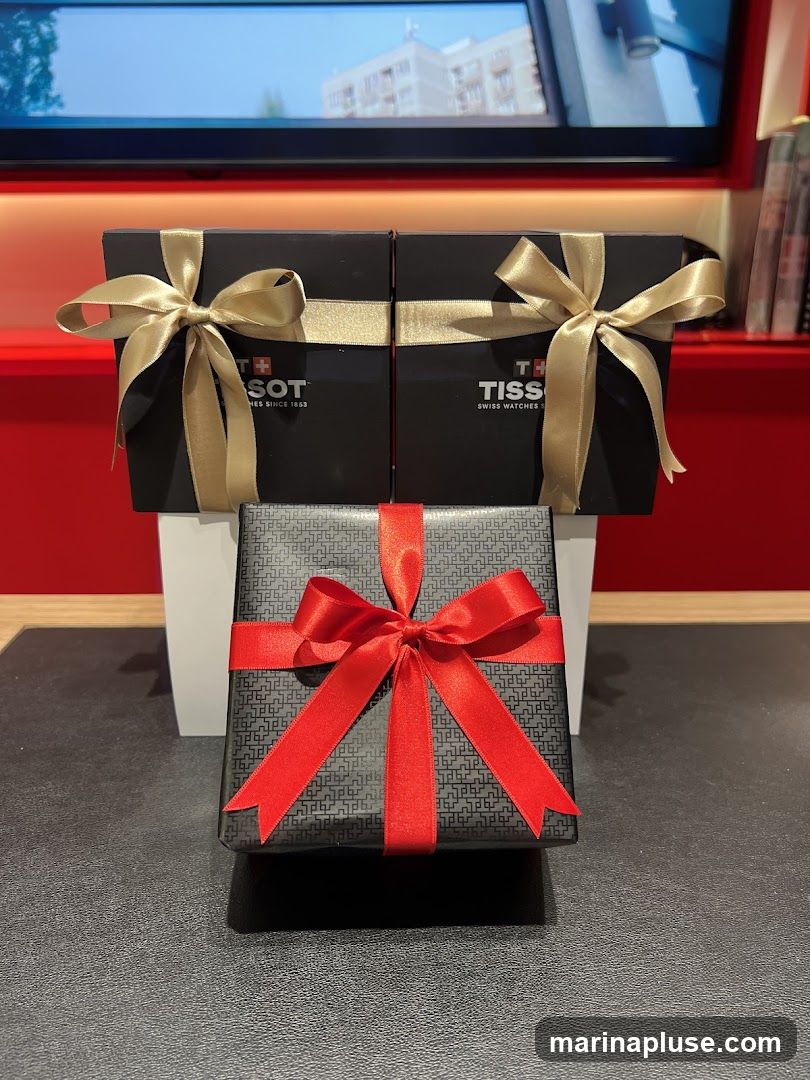 Two black Tissot gift boxes with gold ribbon bows are displayed behind a black gift-wrapped box with a red ribbon bow. The Tissot logo and "SWISS WATCHES SINCE 1853" are visible on the black boxes. A screen displaying a building is in the background.