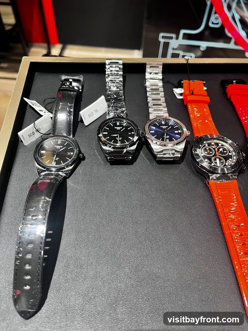 A display of four Tissot watches on a black textured surface. The watches have various strap types, including black leather and metal bracelets, and one has a bright orange strap. Price tags are visible on two of the watches.