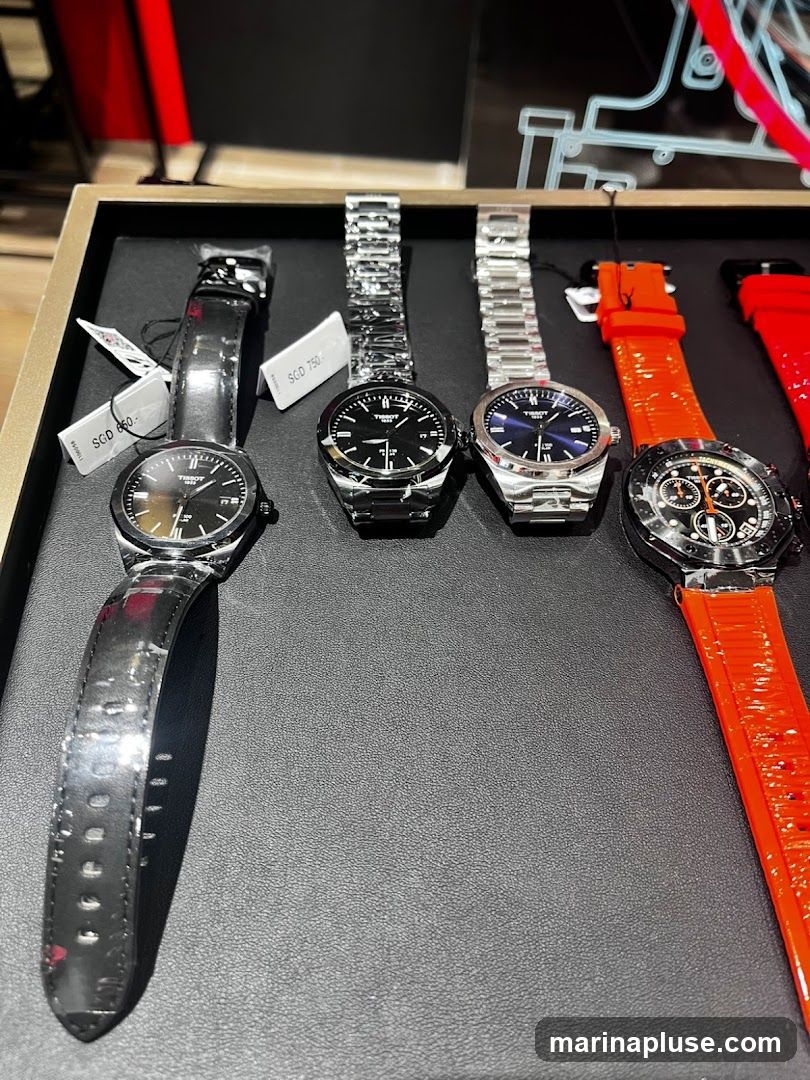 A display of four Tissot watches on a black textured surface. The watches have various strap types, including black leather and metal bracelets, and one has a bright orange strap. Price tags are visible on two of the watches.