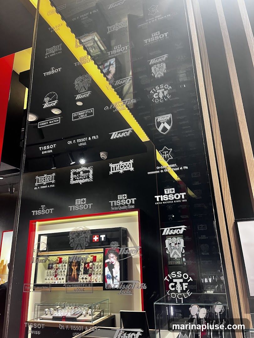 A display case filled with Tissot watches, with the Tissot logo and branding visible on the wall behind. The display features multiple watches in glass cases and on shelves, with some promotional images of models wearing watches. The wall is adorned with various Tissot logos and historical branding.