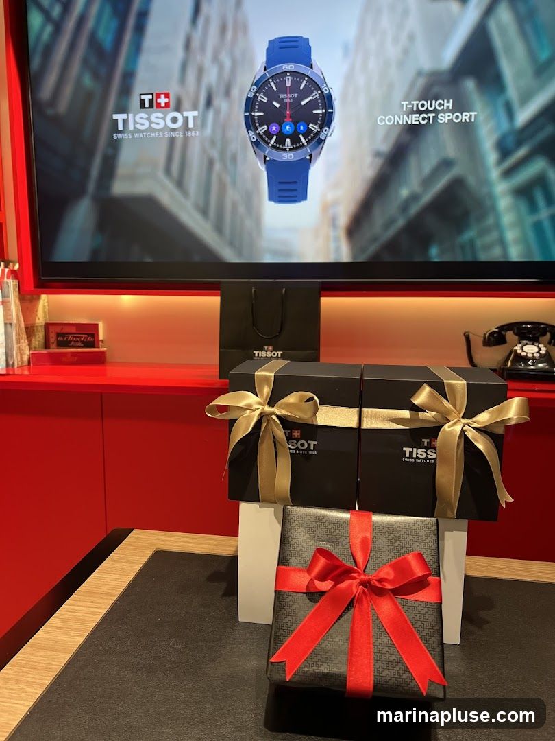 A display featuring a large screen showing an advertisement for a Tissot T-Touch Connect Sport watch. Below the screen are several gift-wrapped boxes with Tissot branding and ribbons, along with a Tissot shopping bag. The background is a bright red shelf.