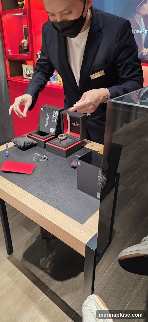 A person wearing a dark suit and a black face mask is presenting a Tissot watch. The watch is in a black and red Tissot branded box, open on a wooden table. Another Tissot branded box is open next to it, and a watch bracelet is displayed. Behind the table, a red display cabinet with Tissot branding is visible, showcasing watches. A Tissot name tag is pinned to the person's suit jacket.