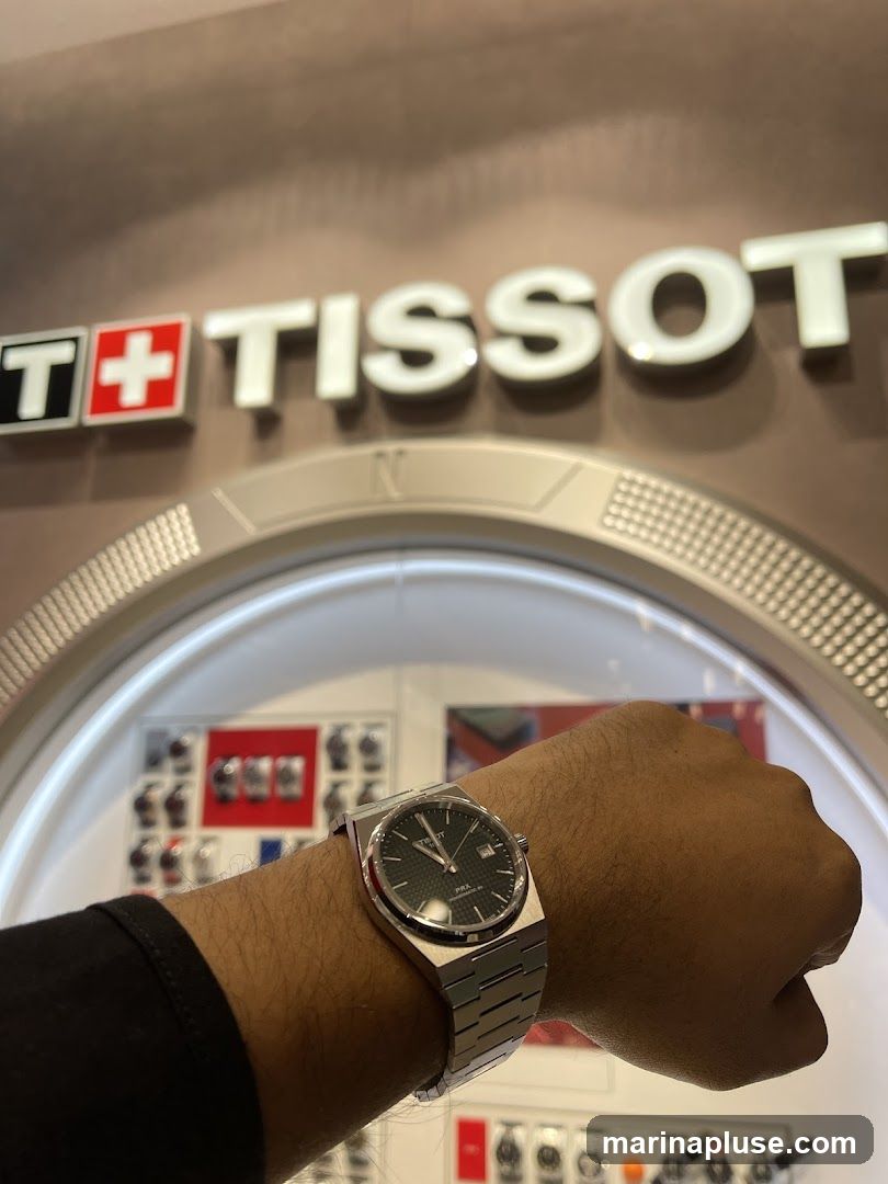 A person's wrist wearing a Tissot PRX watch is shown in the foreground. Behind the wrist, a display case with various Tissot watches is visible. The Tissot logo is prominently displayed above the display case.