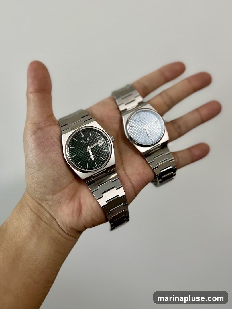 A hand holds two Tissot watches against a plain background. The watch on the left has a dark green dial, and the watch on the right has a light blue dial. Both watches have stainless steel bands and cases.
