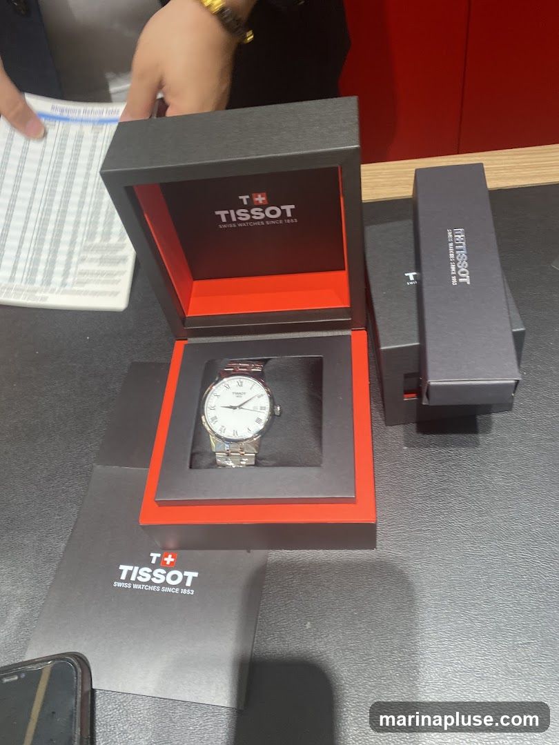 A Tissot watch is displayed in its open black and red presentation box. The watch has a white face with Roman numerals and a silver metal band. Next to the open box is a closed Tissot watch box. A Tissot branded pamphlet or card is also visible. In the background, a person's hand is visible holding a document with text and columns.