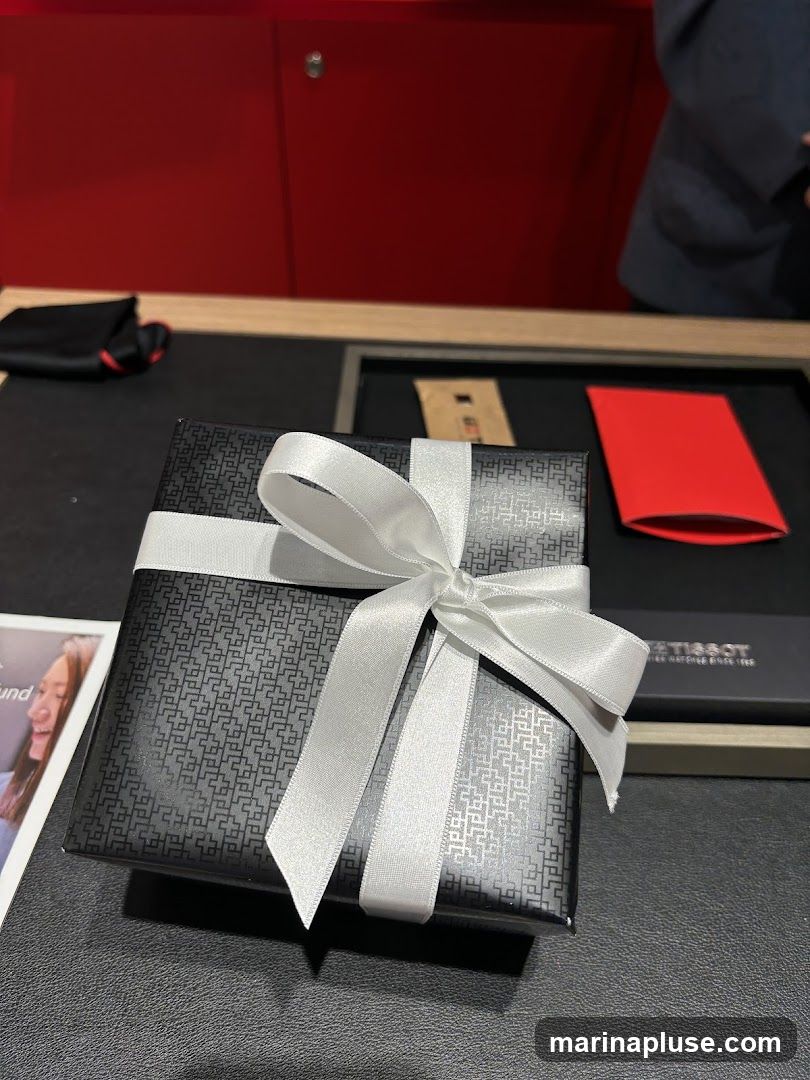 A black gift box with a white ribbon is placed on a dark surface. The box has a textured pattern. In the background, there is a red cabinet and a person wearing a dark jacket. A red envelope and a small brown package are also visible on the surface. A Tissot watch box is to the right of the gift box.
