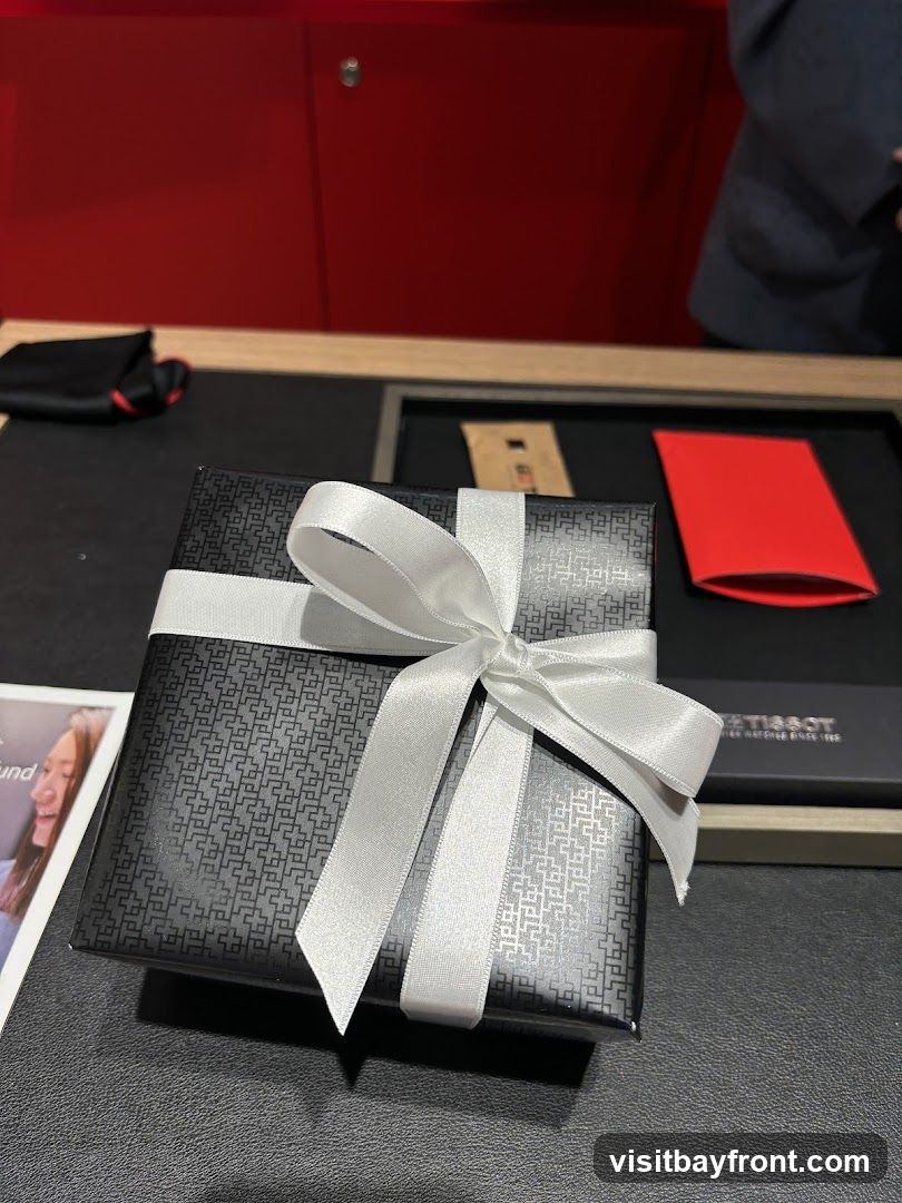 A black gift box with a white ribbon is placed on a dark surface. The box has a textured pattern. In the background, there is a red cabinet and a person wearing a dark jacket. A red envelope and a small brown package are also visible on the surface. A Tissot watch box is to the right of the gift box.