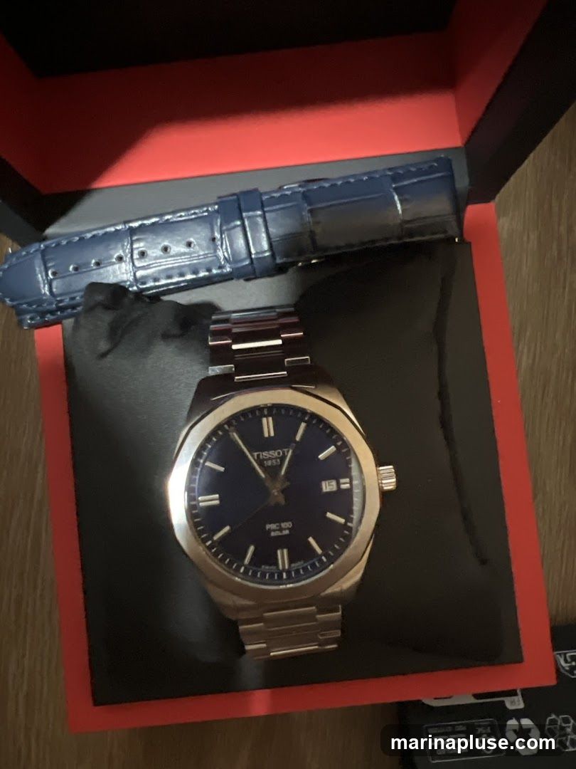 A Tissot PRC 100 Solar watch with a blue dial and a stainless steel bracelet is displayed in its open box. A dark blue, textured watch strap is placed above the watch. The box has red trim.