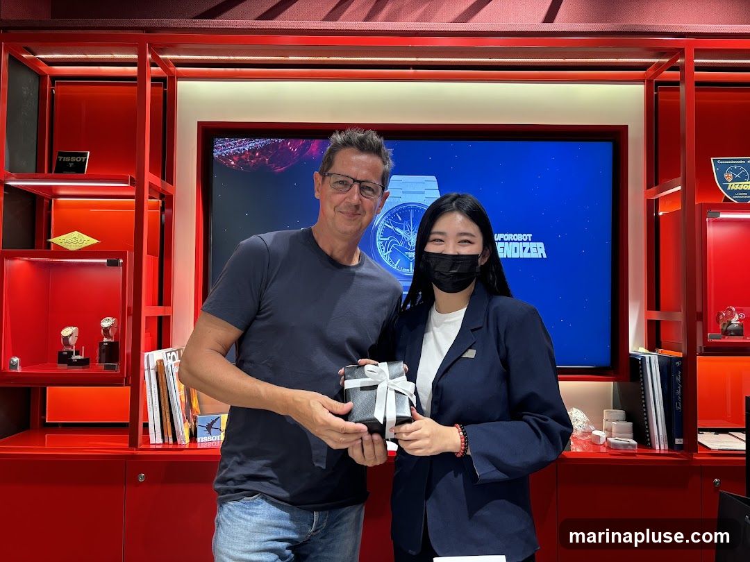 A man and a woman are standing in a Tissot watch store. The man is wearing a dark blue t-shirt and jeans, and the woman is wearing a dark blue blazer over a white shirt. They are both smiling and holding a gift-wrapped box with a white ribbon. The store has red shelves displaying watches and Tissot branding. A large screen in the background shows a watch face and text that reads "UPOROBOT ENDIZER".