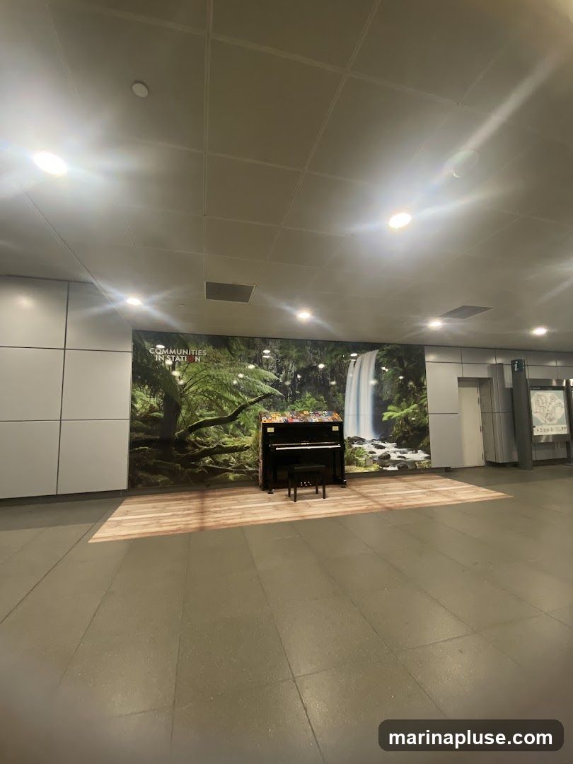 A black upright piano with a bench in front of it is placed in a public area. Behind the piano is a large mural depicting a waterfall and lush greenery. The floor in front of the mural has a wood plank design.