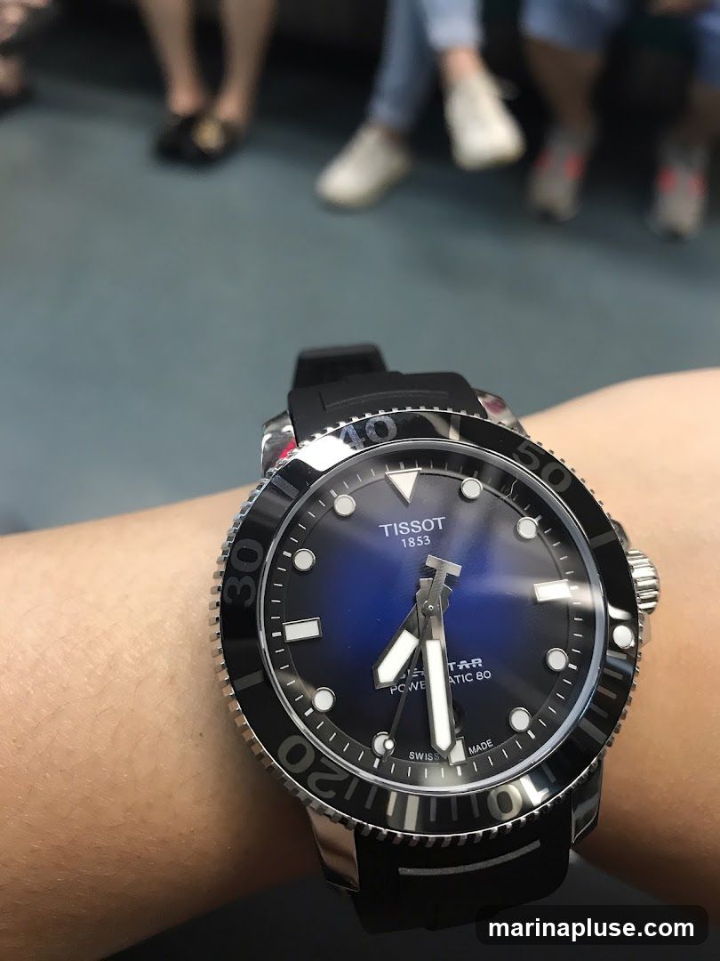 A close-up shot of a Tissot watch on a person's wrist. The watch has a blue dial with silver hands and markers. The bezel is black with white numerals. In the background, out of focus, are the legs of several people sitting down.
