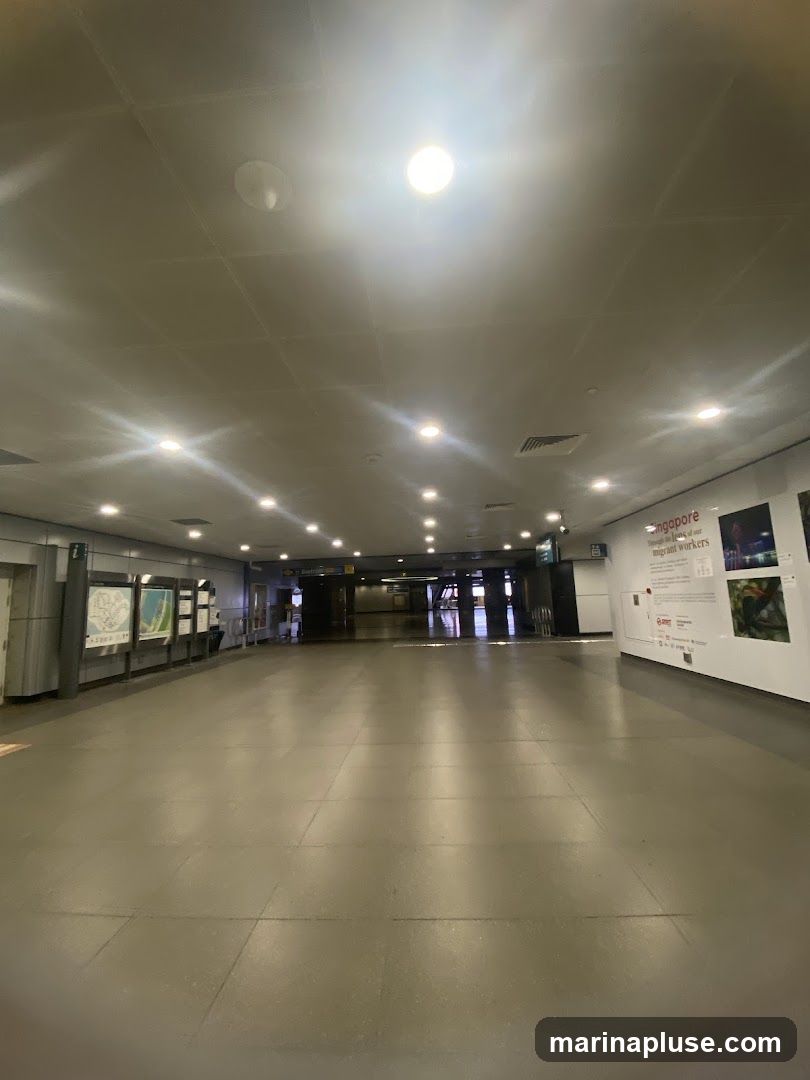 An empty, well-lit hallway with a tiled floor and a drop ceiling. On the left wall, there are several display boards with maps or information. On the right wall, there are large posters, one of which has text that includes "Singapore" and "migrant workers". The hallway extends into the distance, with more lights visible further down.