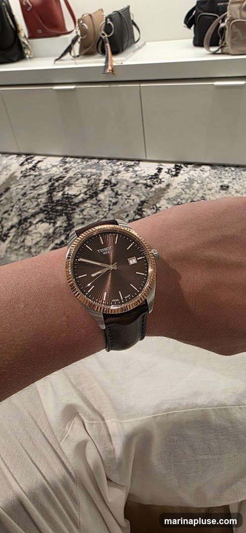 A close-up shot of a Tissot watch with a brown dial and rose gold bezel worn on a person's wrist. The watch has a leather strap. In the blurred background, several handbags are displayed on a shelf.