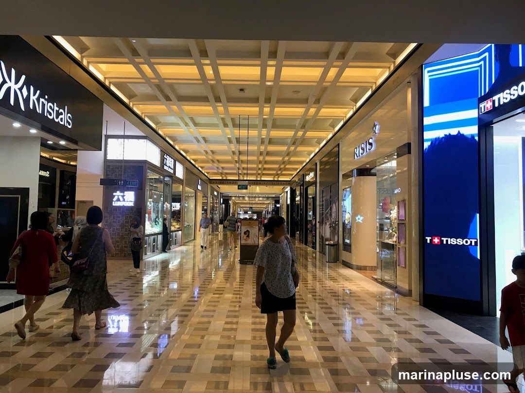 A view down a brightly lit shopping mall corridor with various storefronts visible on both sides. People are walking through the mall. Store signs include Kristals, RISIS, and Tissot. The ceiling features a grid-like illuminated design.