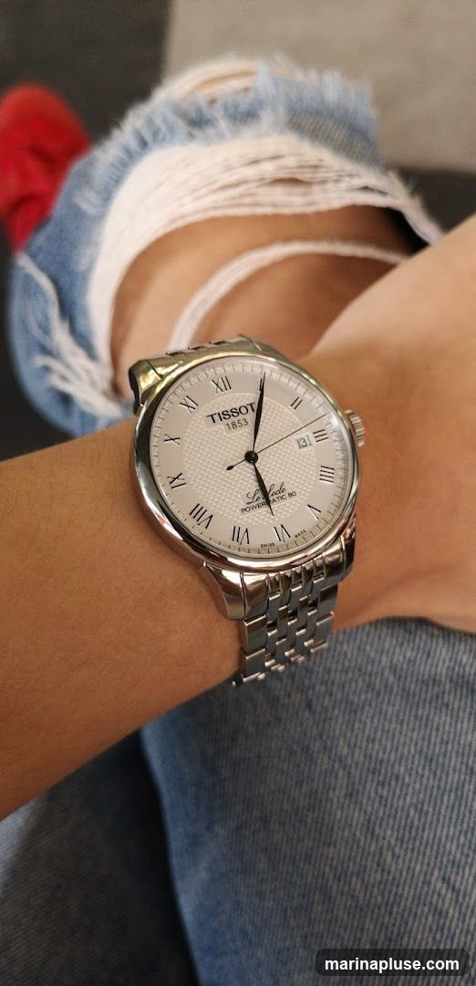 A close-up shot of a Tissot watch on a person's wrist. The watch has a silver band and a white face with Roman numerals. The brand name "TISSOT 1853" is visible at the top of the face, and "Le Locle POWERMATIC 80" is written below the hands. The person is wearing ripped blue jeans and a red sock.