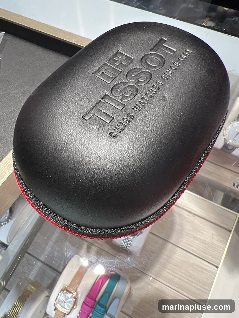A black Tissot watch case with the Tissot logo and "SWISS WATCHES SINCE 1853" embossed on it. Below the case, a display case shows several watches, including one with a square face and a brown strap, and others with colorful straps.