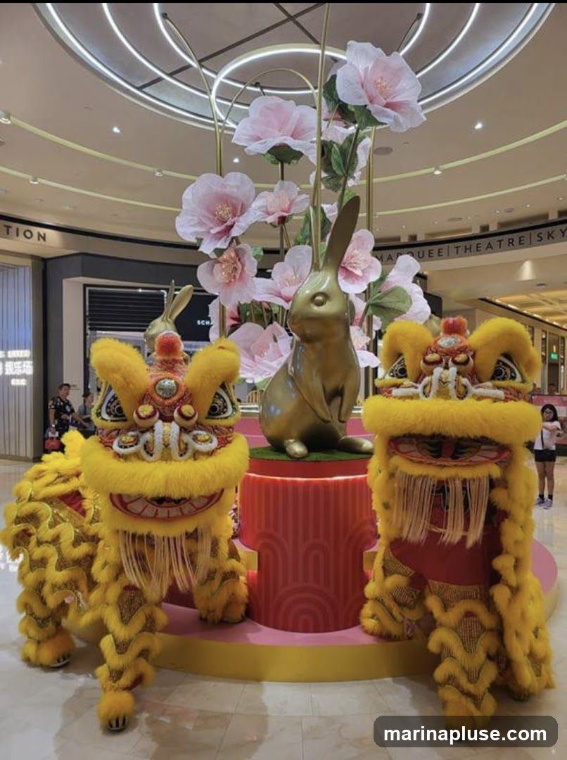 A festive display featuring golden rabbit statues and lion dance costumes, adorned with large pink paper flowers, likely for Lunar New Year celebrations in a shopping mall.