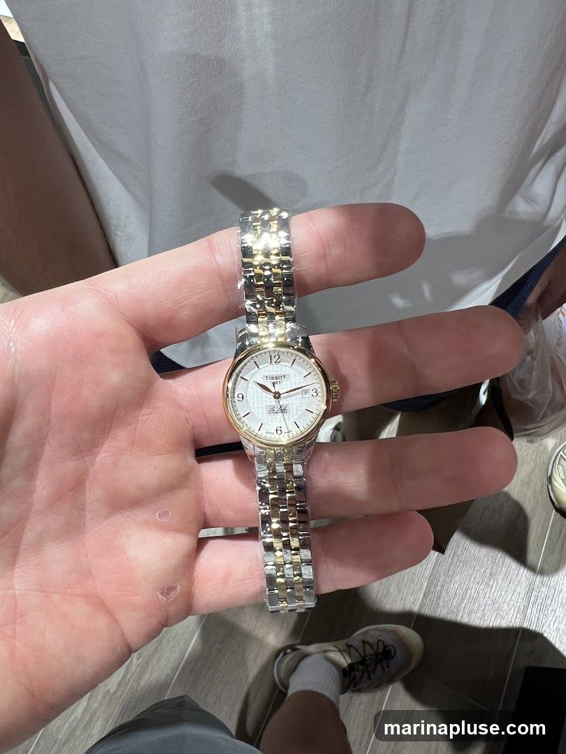 A person is holding a Tissot watch with a two-tone metal band. The watch face is white with gold accents and Roman numerals. The background shows a person wearing a white t-shirt and grey shorts, with sneakers visible at the bottom.