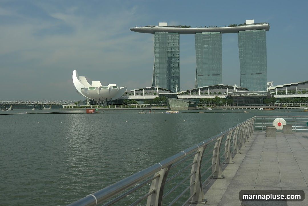 A wide shot of the Marina Bay Sands hotel and the ArtScience Museum in Singapore, viewed across a body of water. The iconic three-tower hotel with its sky park is in the background, while the lotus-shaped museum is to the left. A walkway with a metal railing is in the foreground.