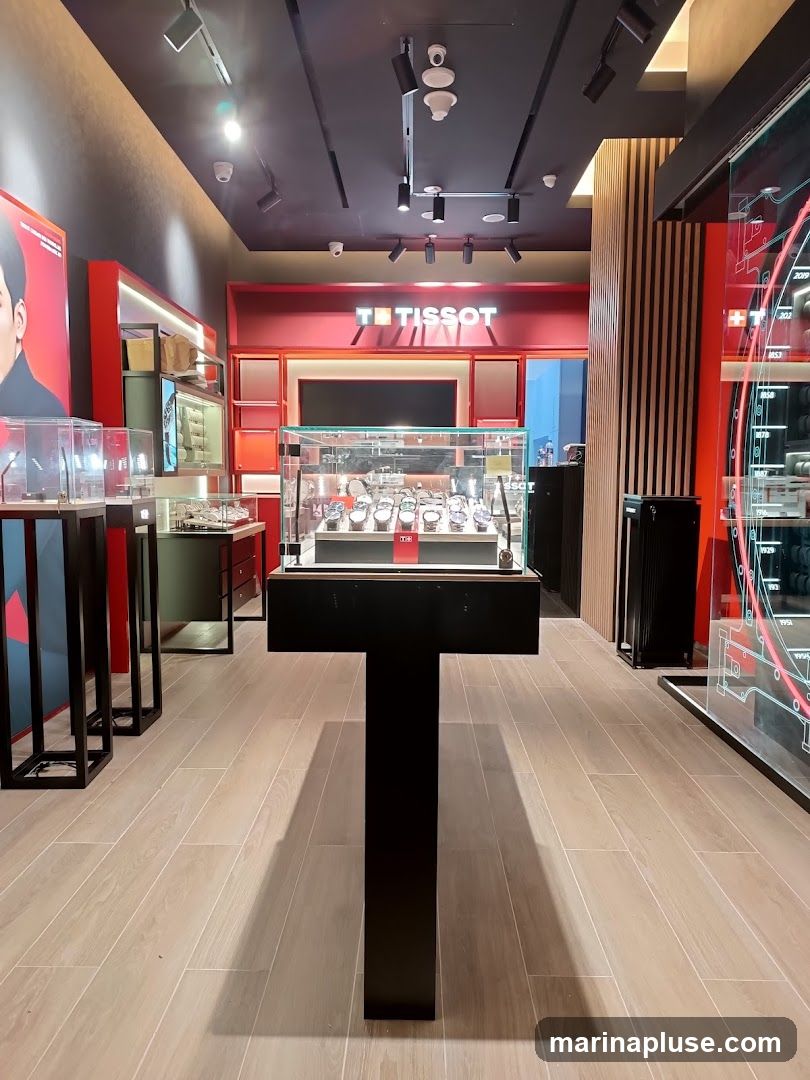 The interior of a Tissot watch store, featuring display cases filled with watches. The store has a modern design with red accents and wooden flooring. A large Tissot logo is visible on the back wall.