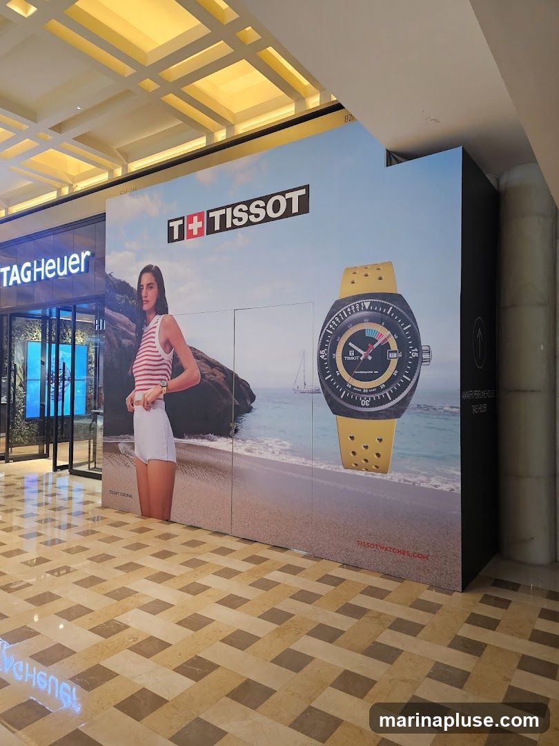 A large advertisement for Tissot watches is displayed on a wall. The advertisement features a woman in casual attire standing on a beach, with a large image of a Tissot watch with a yellow strap. The Tissot logo is prominently displayed at the top. To the left, a TAG Heuer store is visible.
