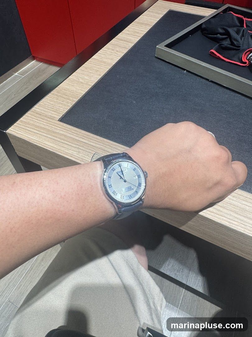 A person's arm with a watch on their wrist is shown. The watch has a silver face with Roman numerals and a blue leather strap. The background appears to be a display counter in a store, with a black textured surface and a wooden trim. A red wall is visible in the upper left corner.