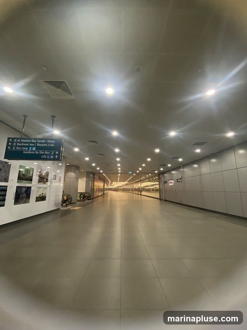 An indoor walkway with a tiled floor and a ceiling with numerous lights. Directional signs indicate routes to Marina Bay Sands, Gardens by the Bay, and a bus stop. Artwork is displayed on a wall to the left. The walkway extends into the distance, with shops or other establishments visible on the right side.