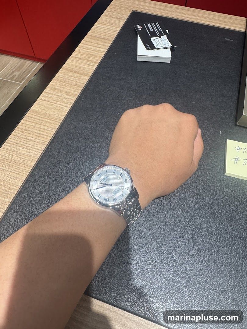 A person's arm wearing a silver Tissot watch with a white patterned dial and Roman numerals. The watch is on a metal link bracelet. The arm is resting on a black textured surface, possibly a desk or counter. In the background, there is a stack of white cards with a black Tissot tag on top. A red cabinet is visible on the left side of the image.