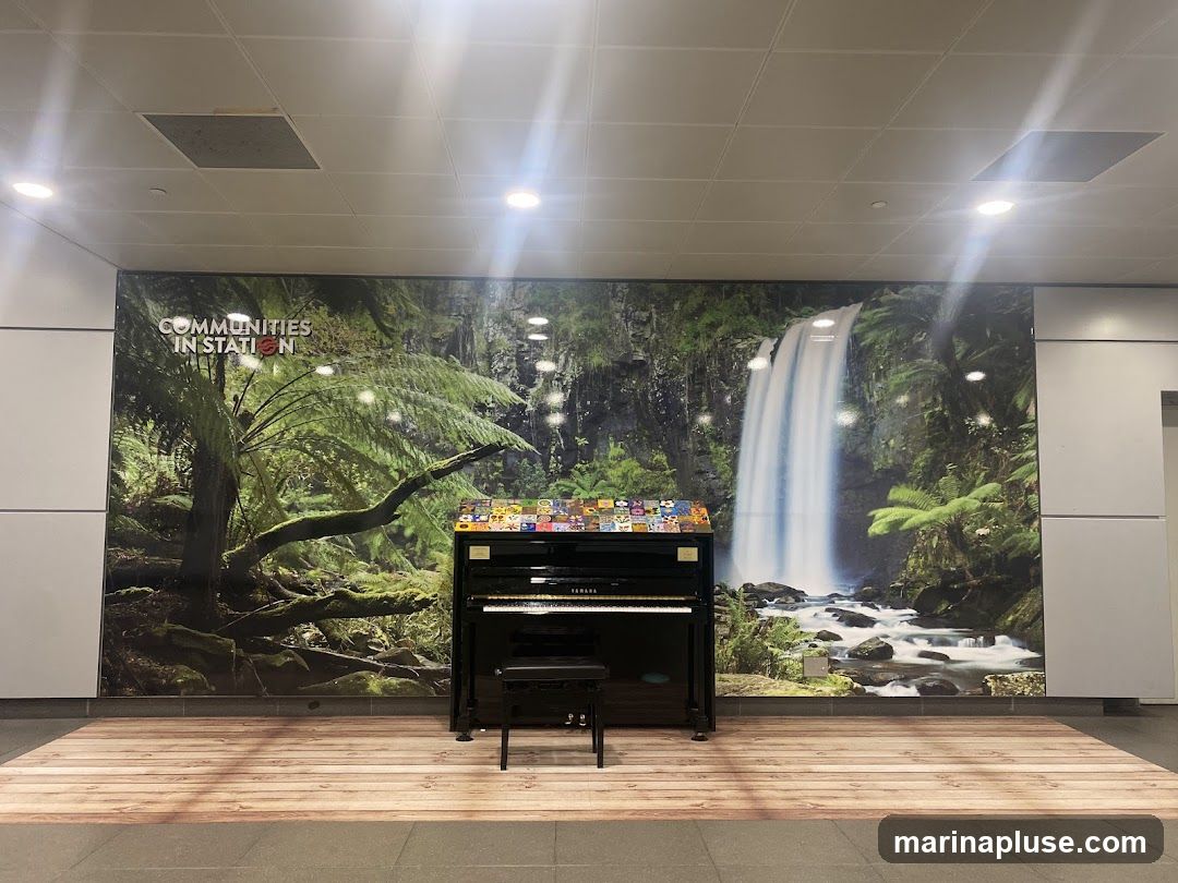 A black upright piano with a bench in front of a large mural depicting a lush forest with a waterfall. The mural has text that reads "COMMUNITIES IN STATION". The floor in front of the piano is designed to look like wooden planks.