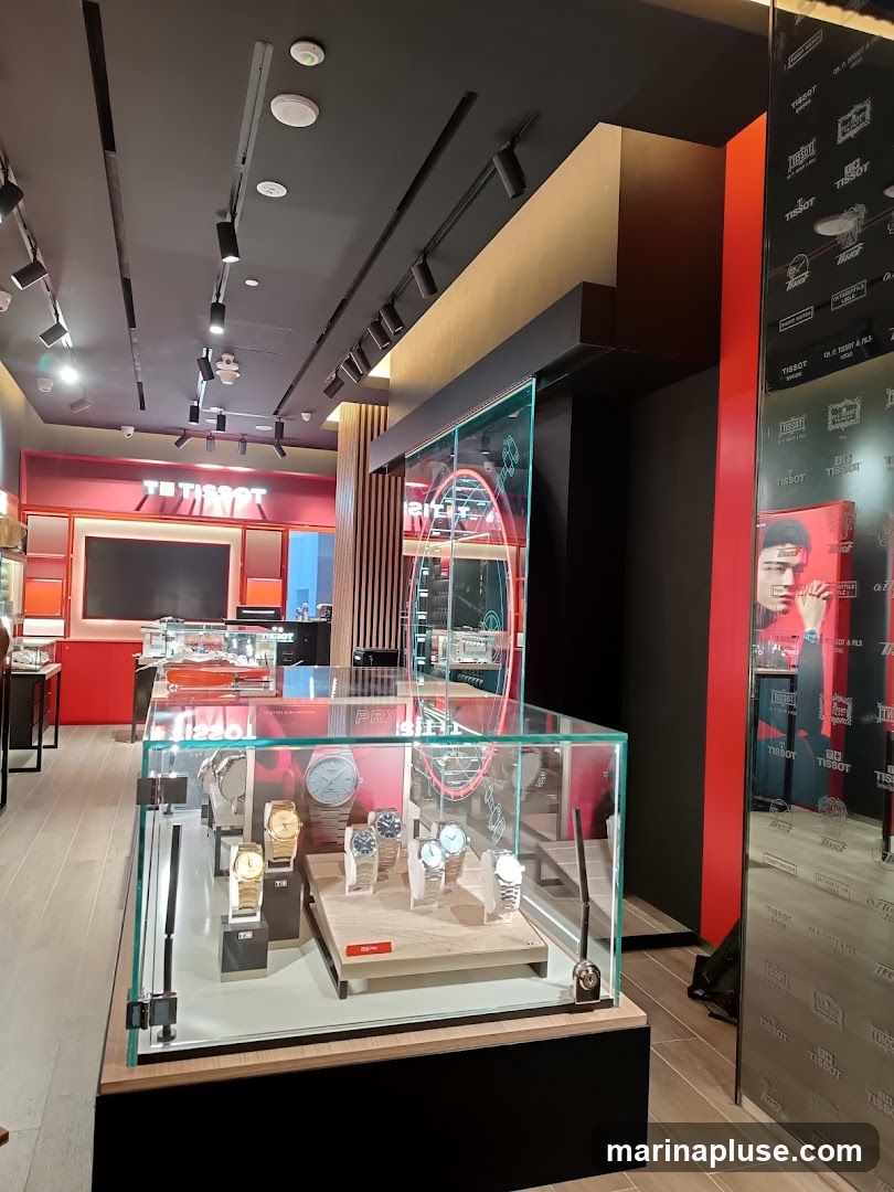 A well-lit interior of a Tissot watch store. In the foreground, a glass display case showcases several Tissot watches on wooden stands. Behind the display, a large glass panel features a neon Tissot logo and a promotional image of a person. The back wall is painted red with Tissot branding and logos visible. The store has a modern design with track lighting on the ceiling and wooden flooring.