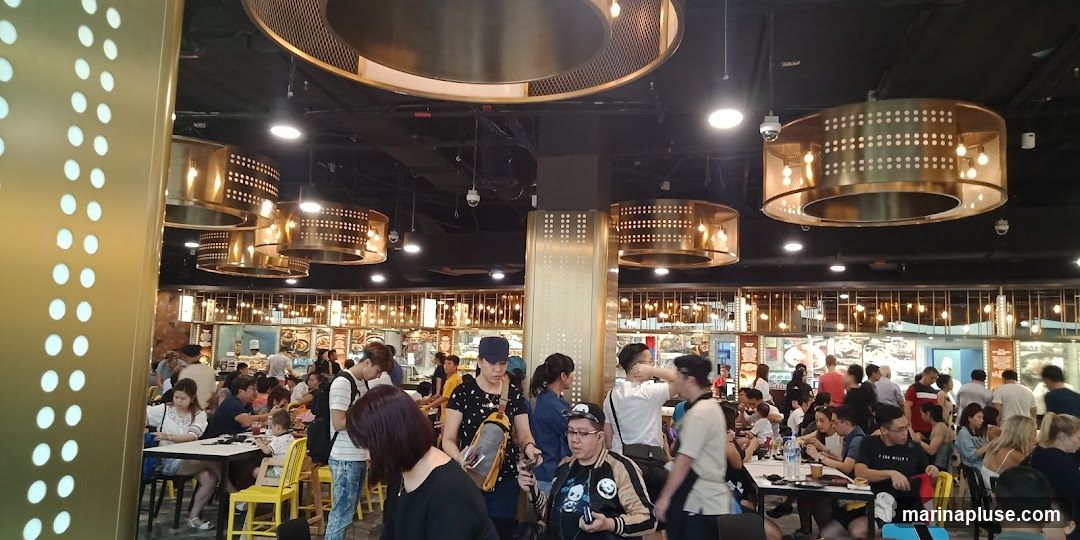 A bustling food court with many patrons seated at tables and ordering food. The ceiling features decorative golden light fixtures. The background shows various food stalls with menus and displays.