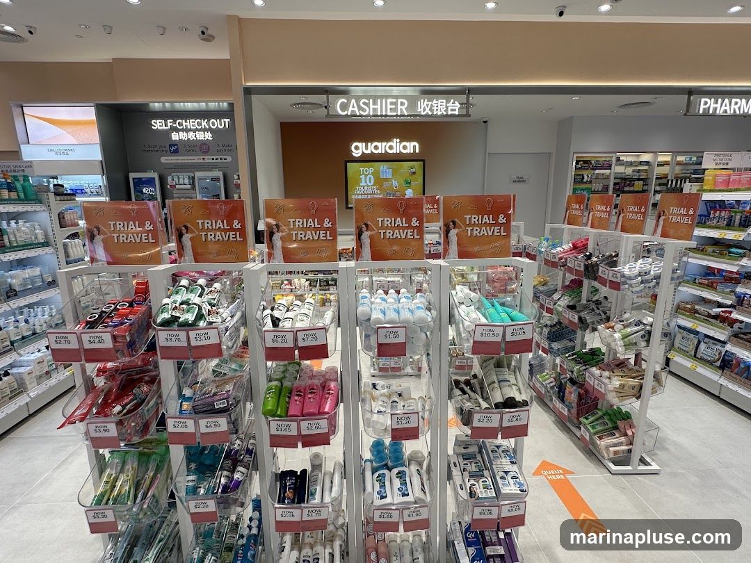A Guardian pharmacy store interior with a prominent display of 'Trial & Travel' sized products. Shelves are stocked with various toiletries, skincare, and travel-sized items. Signs indicate 'Self-Check Out' and 'Cashier'. An orange arrow on the floor indicates a queue direction.