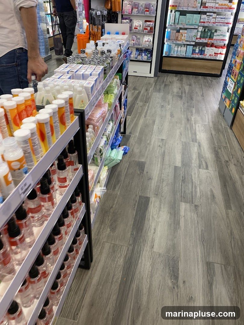 A view down an aisle in a store, showcasing shelves stocked with various products. On the left, shelves are filled with rows of clear bottles with red spray tops, likely personal care items. Further back, more shelves display boxes and bottles of what appear to be health and beauty products, including baby care items and skincare. In the background, a person is walking away, and more shelves with packaged goods are visible. The flooring is a grey wood-look laminate.