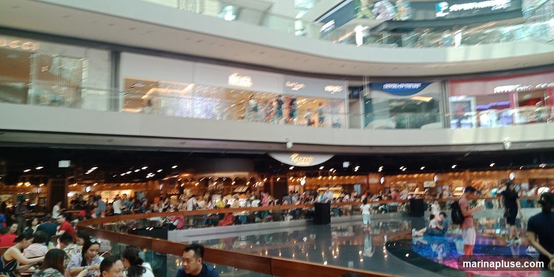 The image shows a bustling interior of a multi-level shopping mall. On the upper levels, various shop fronts are visible, including what appears to be a Gucci store and other retail outlets. Below, a lively food court area is filled with people dining at tables. The architecture features modern design with reflective surfaces and bright lighting.
