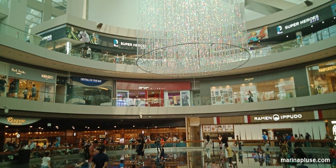 An interior view of a multi-level shopping mall atrium, featuring a large, sparkling chandelier. Various shops are visible on different levels, including "SUPER HEROES CAFE", "Paul Smith Junior", "KENZO", "DENTAL ON THE BAY", "RAMEN IPPUDO", and "EMPORIO". People are seen browsing and walking around the mall. The lower level appears to be a food court or dining area with many patrons.