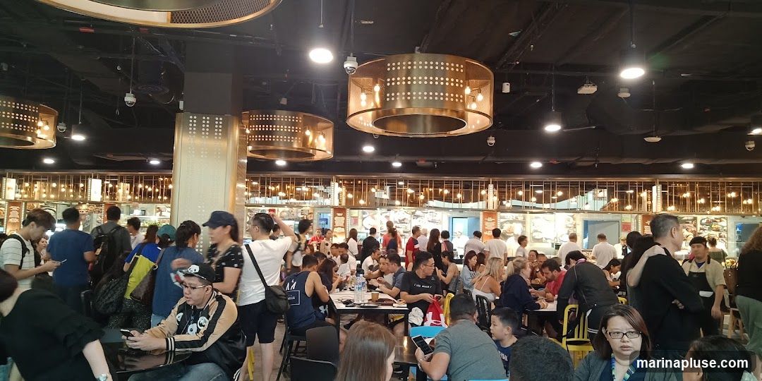 A crowded food court with many people dining and queuing. The ceiling features modern, circular golden light fixtures. The background shows various food stalls with menus displayed.