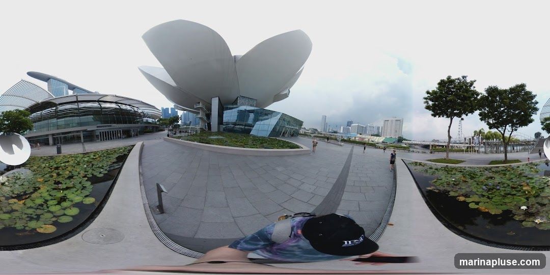 A panoramic view of the ArtScience Museum, with its distinctive lotus-inspired architecture, and the Marina Bay Sands hotel in the background. The scene includes a plaza with paving stones, a pond with lily pads, and trees. Several people are visible walking around the area. The sky is overcast.