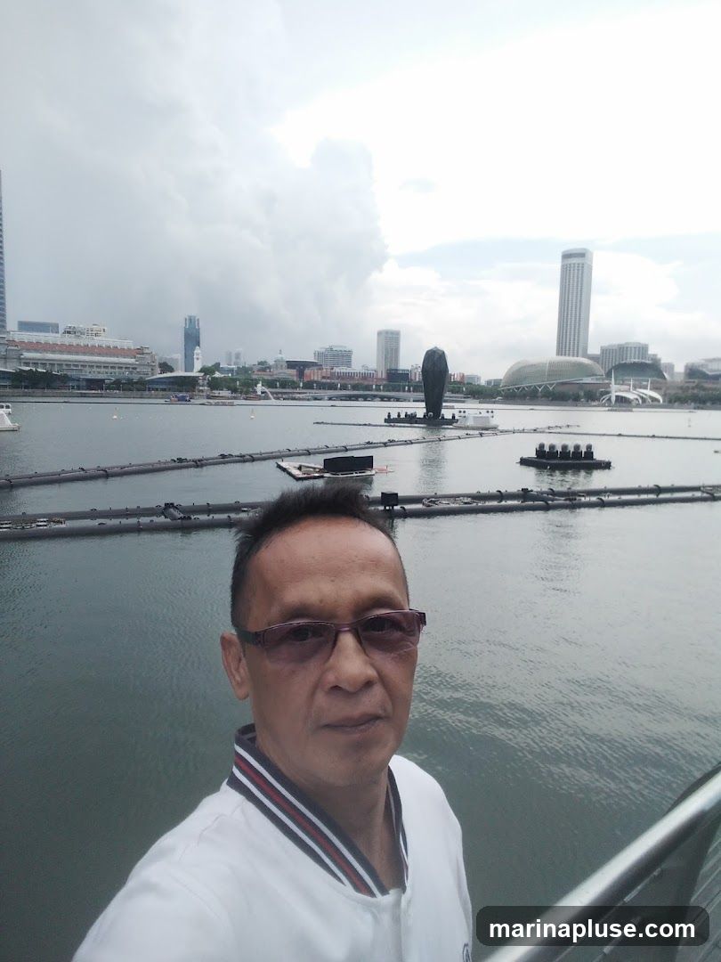 A man wearing glasses and a white jacket with red and blue trim takes a selfie in front of a body of water. In the background, a cityscape with modern buildings and a cloudy sky is visible. There are also some floating structures in the water.