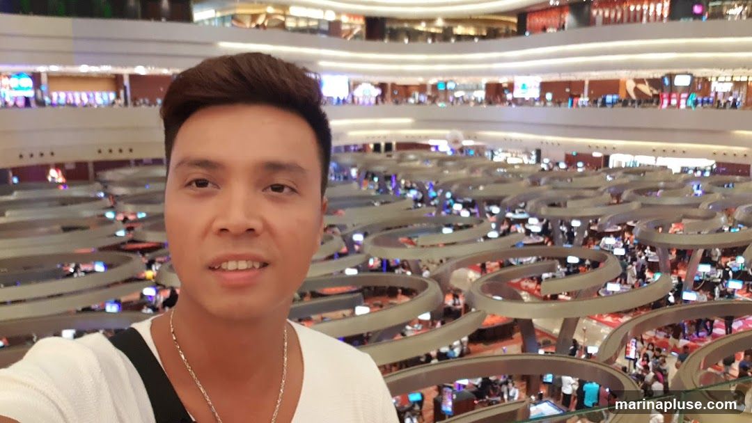 A man takes a selfie in a large, multi-level casino. The background shows rows of gaming tables and slot machines with many people playing. The casino appears to be brightly lit with various colorful lights.
