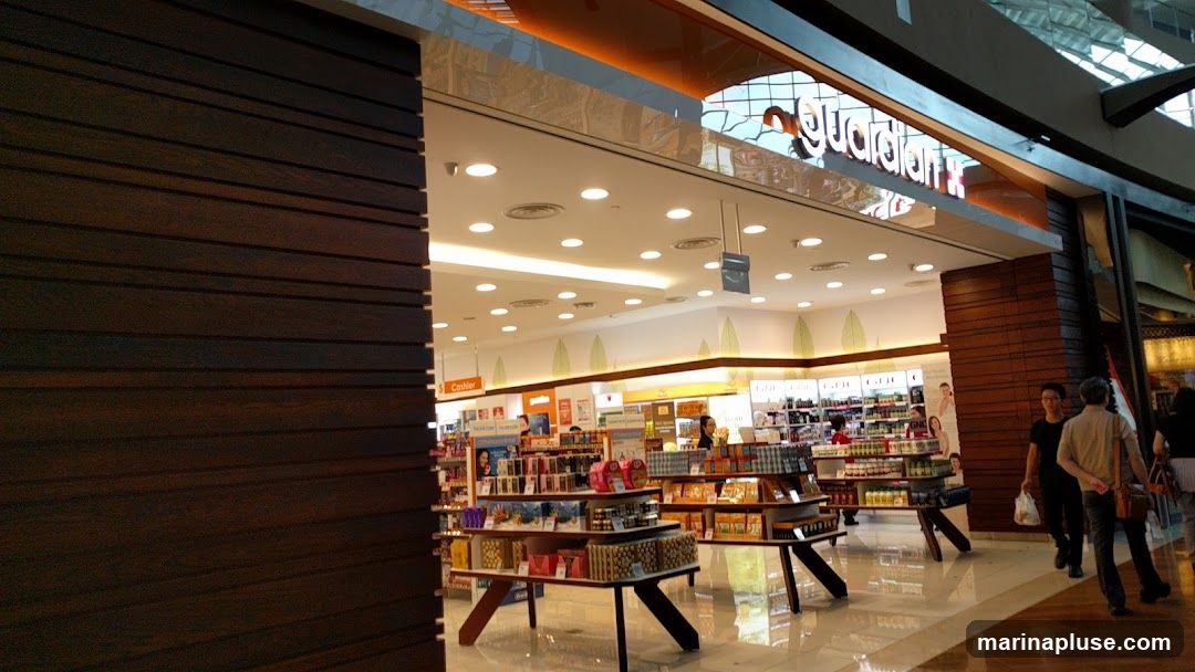 The exterior of a Guardian store, with its prominent signage visible above the entrance. The store is well-lit and stocked with various health and beauty products displayed on shelves and stands. Several customers are seen browsing inside and walking past the entrance. The interior features a clean and modern design with wooden accents.