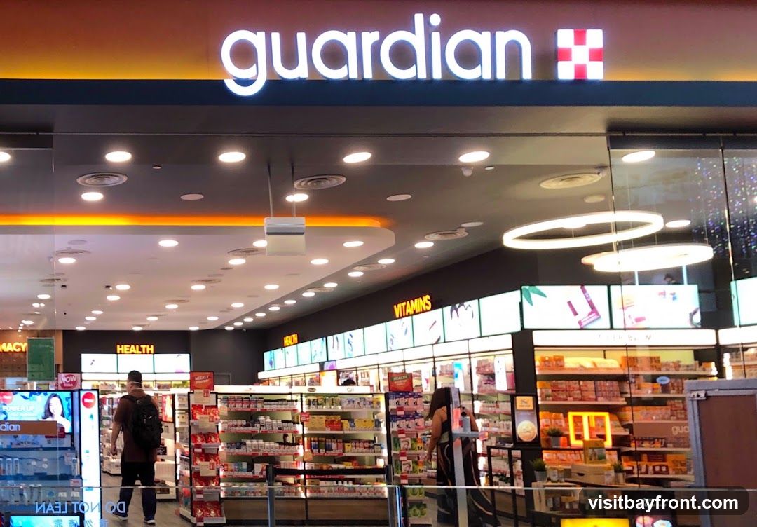 The exterior of a Guardian pharmacy with its prominent illuminated logo above the entrance. Inside, shelves are stocked with health and beauty products, and customers are browsing.