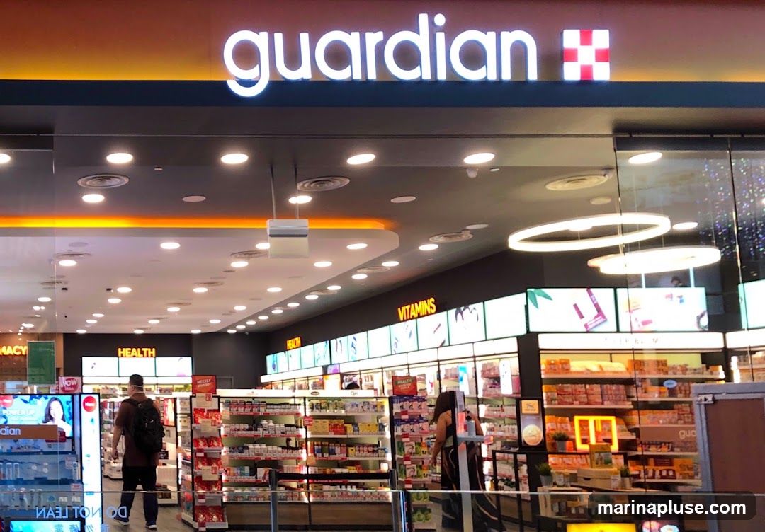 The exterior of a Guardian pharmacy with its prominent illuminated logo above the entrance. Inside, shelves are stocked with health and beauty products, and customers are browsing.