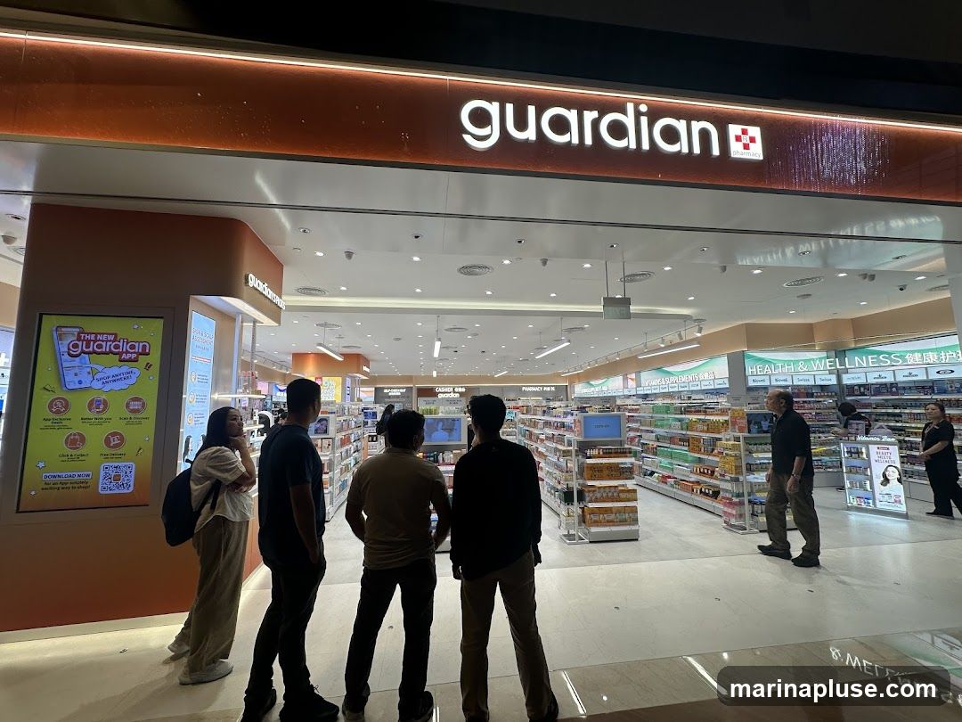 A Guardian pharmacy storefront with a large illuminated sign above the entrance. Inside, shelves are stocked with various health and wellness products. Several people are browsing the aisles.