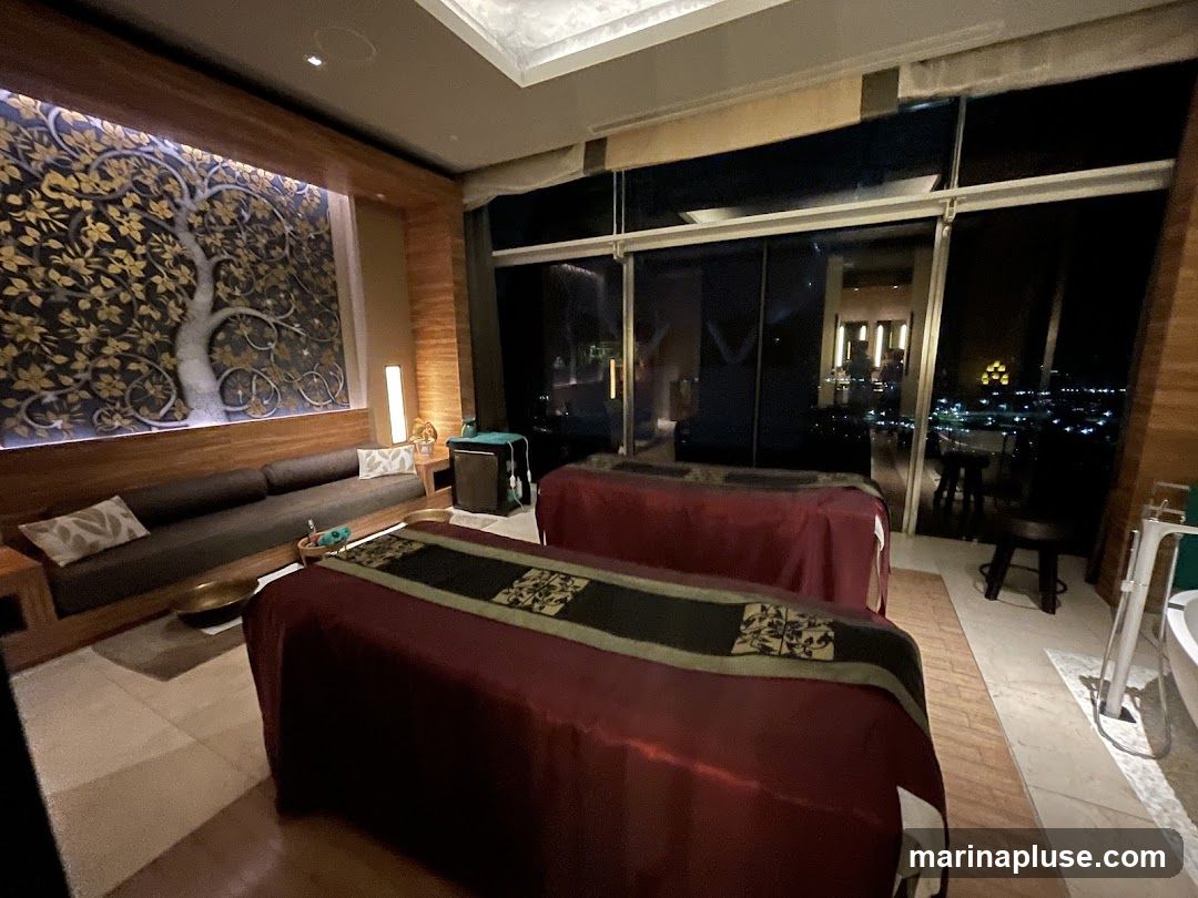 A luxurious spa room with two massage beds, a long sofa, and a large window overlooking a city at night. The room features a decorative wall panel with a tree motif and warm lighting.