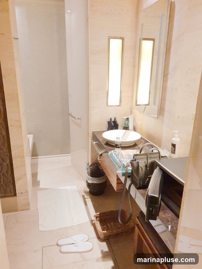 A well-lit bathroom vanity area with a sink, mirror, and toiletries. A shower stall is visible to the left. White slippers and a bath mat are on the floor. A woven basket and a trash can are near the vanity.