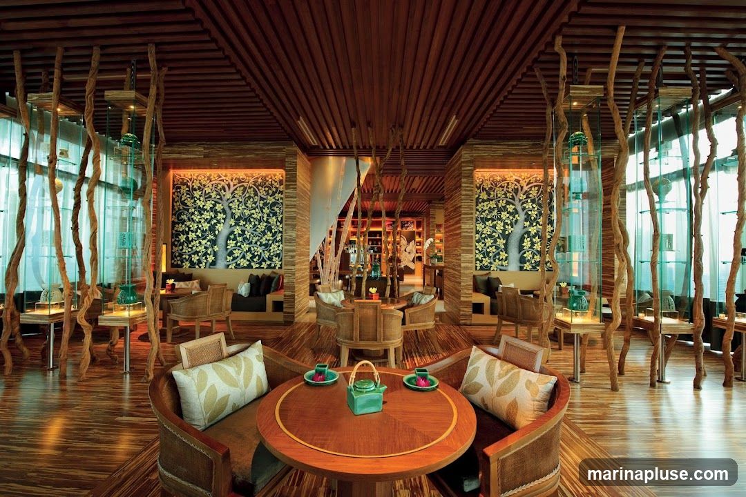 A beautifully designed lounge area with a natural, organic theme. The space features wooden floors and ceilings, with decorative wooden branches creating a unique visual element. Two large, framed artworks depicting stylized trees with golden leaves adorn the walls. Seating includes comfortable chairs and sofas arranged around round wooden tables. A green teapot and cups are placed on one of the tables, suggesting a tea service. Glass display cases with decorative items are visible in the background.