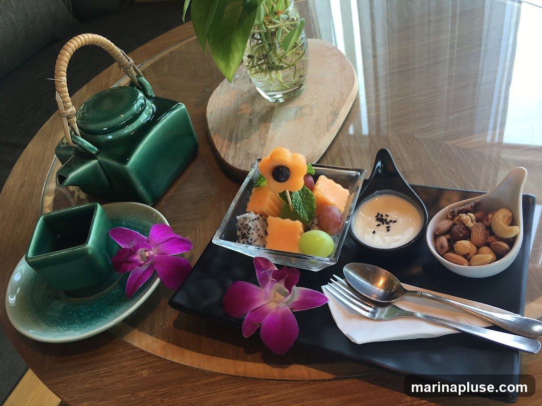 A dark green teapot and two small square cups sit on a saucer. A platter holds a square glass dish of fruit salad, a small black dish with a white dip topped with black seeds, and a white dish of mixed nuts. Two purple orchids are placed on the platter. A fork and spoon rest on a white napkin.
