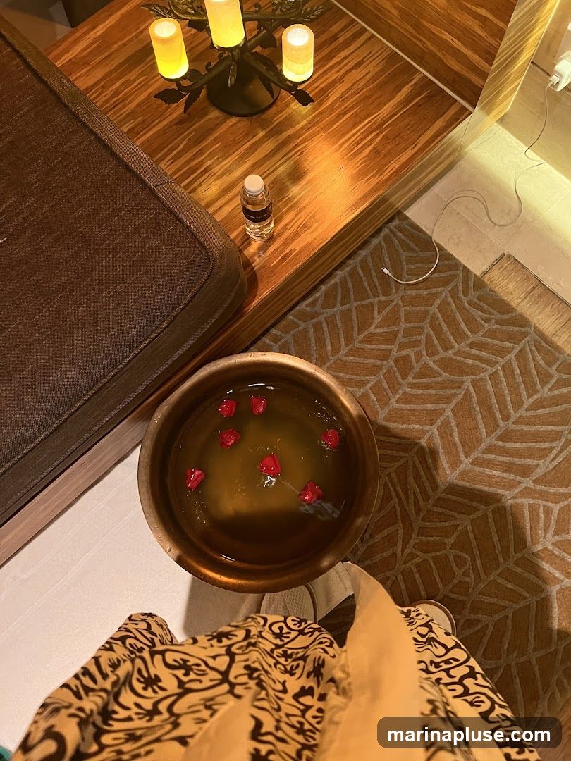 A close-up overhead view of a spa setting. In the foreground, a person's legs are visible, wearing patterned loungewear. A copper bowl filled with water and floating red flower petals sits on a textured mat. To the left, a dark brown cushioned bench is partially visible. On a wooden table behind the bench, there is a small bottle of liquid and a decorative candelabra with three lit candles. A white charging cable is visible on the floor to the right.