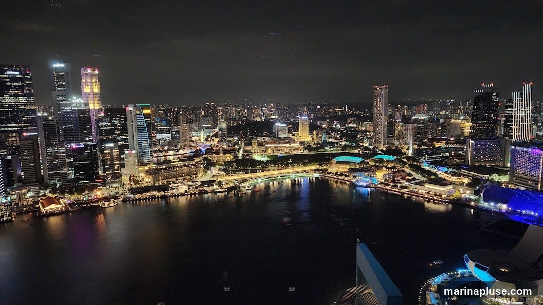 A panoramic view of a city skyline at night, with numerous skyscrapers illuminated by lights. A body of water, likely a river or bay, is in the foreground, reflecting the city lights. Several boats are visible on the water. The cityscape includes modern architecture and some older buildings, with a bridge spanning the water. The sky is dark and cloudy.