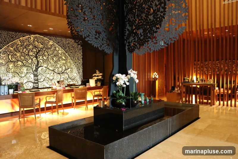 A hotel lobby with a decorative tree sculpture and a reception desk. The reception desk has a large, ornate tree mural behind it. There are chairs at the desk and a water feature in the foreground. The background features wooden paneling and shelves with various items.